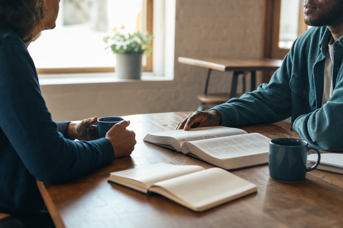Two people having a calm, supportive conversation at a quiet café table with open books and warm natural light
